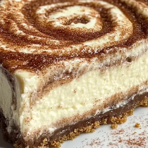 A close-up view of a slice of cinnamon roll cheesecake with swirls of cinnamon sugar, topped with cream cheese frosting, served on a white plate.