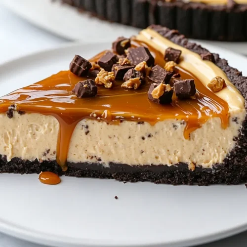 Close-up view of a slice of Reese's Caramel Cheesecake with creamy filling, crunchy Oreo crust, caramel drizzle, and chocolate ganache on a white ceramic plate.