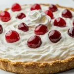 A close-up of a creamy Root Beer Float Pie with a graham cracker crust, topped with whipped cream and maraschino cherries, served on a white plate.