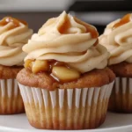 Close-up of apple pie cupcakes with cinnamon buttercream, caramel drizzle, and tender apple filling, served on a white plate with a cozy kitchen backdrop.